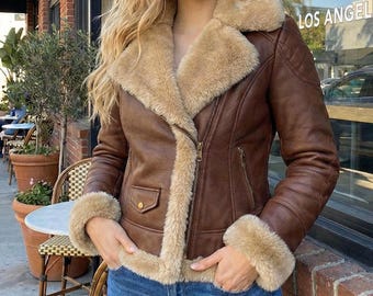 May include: Brown faux leather jacket with a beige shearling lining and trim. The jacket features a zip closure, two front pockets, and quilted shoulder detailing. The cuffs and collar are lined with the same beige shearling material.