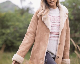 May include: A tan suede coat with a cream-colored shearling lining and trim. The coat has a button-up front and long sleeves. The person in the image is wearing a gray sweater and dark jeans.