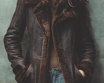 May include: A brown leather coat with a faux fur collar and lining. The coat has a classic design with a button closure and pockets. The model is wearing a brown sweater and blue jeans. The coat appears to be vintage or distressed.