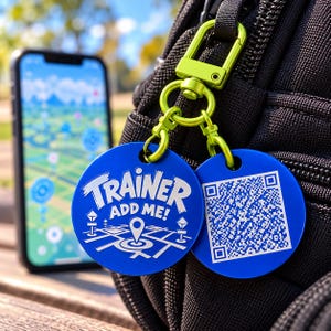 Puede incluir: Dos etiquetas circulares de metal azul unidas a un mosquetón verde lima. Una etiqueta presenta el texto blanco TRAINER ADD ME! sobre un icono de mapa. La segunda etiqueta muestra un código QR blanco. Las etiquetas están sujetas a un bolso negro.