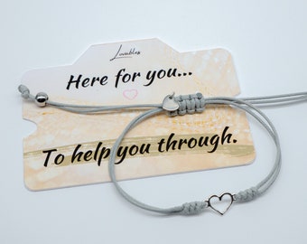 May include: Gray friendship bracelet with a silver heart charm. The bracelet is on a card that says "Here for you... To help you through."