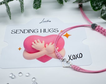 May include: Pink friendship bracelet with a silver heart charm. The bracelet is on a card that says "Sending Hugs" and "XOXO".