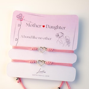 Peut inclure: Deux bracelets d'amitié roses avec des breloques en forme de cœur argentés. La carte indique "Mère Fille Un lien comme aucun autre" et "Lovable Gifts You'll Love".