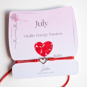May include: Red string bracelet with a silver heart charm. The card says "July, Vitality Energy Passion, Ruby,  Lovable Gifts You'll Love."