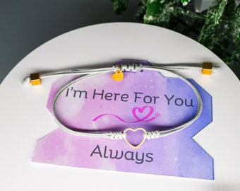 May include: A gray friendship bracelet with a gold heart charm and gold cube accents. The bracelet is displayed on a card with the text "I'm Here For You Always". The card has a purple and pink gradient background.