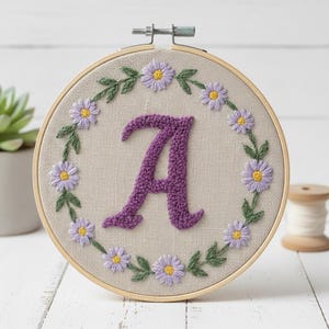 Op de afbeelding: Geborduurde hoepelkunst met een paarse letter "A" in het midden, omringd door een krans van geborduurde madeliefjes en groene bladeren. De achtergrond is beige stof en de hoepel is van hout.