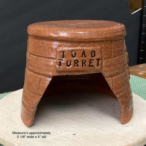 May include: A handmade, brown ceramic toad turret with a brick-like texture. The turret has an arched opening and a flat top. The text "TOAD TURRET" is inscribed on the front. Measures approximately 5 1/8" wide x 4" tall.