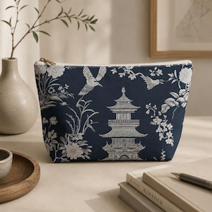 May include: A dark blue zipped pouch featuring a white toile-style print of a multi-tiered pagoda, flying birds, and floral branches. The rectangular bag has a gold-toned metal zip closure along the top edge and a structured base.