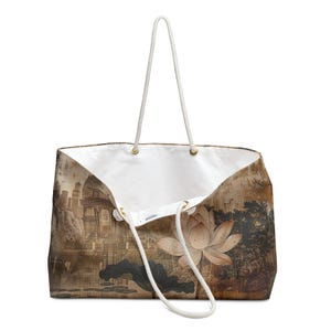 May include: A large tote bag with a white interior and a scenic print. The exterior features a sepia-toned landscape with a lotus flower and a cityscape. The bag has white rope handles and gold-colored eyelets.