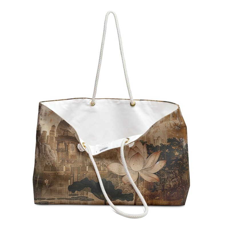May include: A large tote bag with a white interior and a scenic print. The exterior features a sepia-toned landscape with a lotus flower and a cityscape. The bag has white rope handles and gold-colored eyelets.