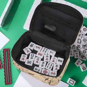 Op de afbeelding: Een open beige Mahjong-draagtas, gevuld met witte tegels met rode en zwarte symbolen. De tas heeft een zwart interieur en staat op een groene ondergrond.