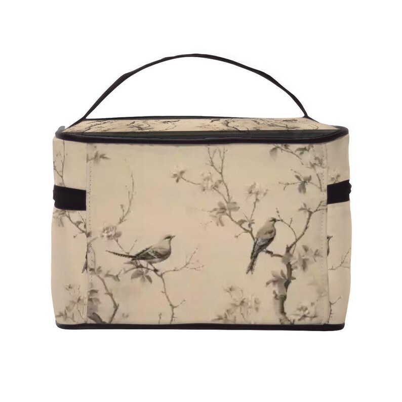Op de afbeelding: Een beige make-uptas met een zwarte handgreep en rand. De tas heeft een bloemen- en vogelontwerp in grijstinten en zwart. De tas heeft een rechthoekige vorm met een handvat aan de bovenkant en een zwarte band aan elke kant.