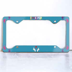 May include: A teal license plate frame with the text "I ❤️ K-POP" in rainbow colors. The frame features hand heart symbols and Korean finger hearts. The frame has a rectangular shape with rounded corners and mounting holes.