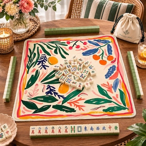 May include: A mahjong set featuring a square play mat with a cream background and a colorful botanical print of green leaves, orange fruits, and red berries. The set includes ivory-colored tiles with traditional symbols and a drawstring storage bag.