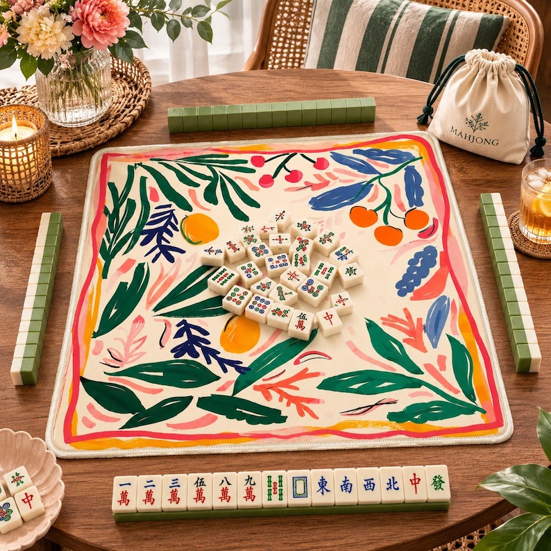 May include: A mahjong set featuring a square play mat with a cream background and a colorful botanical print of green leaves, orange fruits, and red berries. The set includes ivory-colored tiles with traditional symbols and a drawstring storage bag.