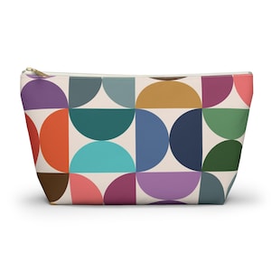 May include: A rectangular cosmetic bag with a vibrant geometric pattern. The design features semi-circles in various colours, including orange, purple, teal, and blue, set against a cream background. The bag has a gold zip.