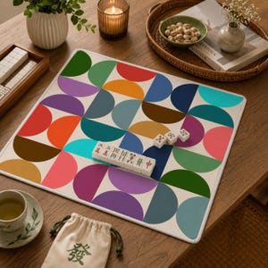 Pu&ograve; includere: Un tappetino da mahjong quadrato con un colorato motivo geometrico di semicerchi nelle tonalit&agrave; del rosso, arancione, giallo, verde, blu, viola e rosa. Diverse tessere bianche da mahjong con caratteri neri e rossi sono disposte sulla superficie.