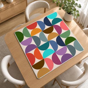 Pu&ograve; includere: Un tappetino quadrato con un motivo geometrico ripetitivo di semicerchi multicolori disposti a griglia. Il design include sfumature di rosso, arancione, giallo, verde, blu, viola e marrone su uno sfondo color crema chiaro.