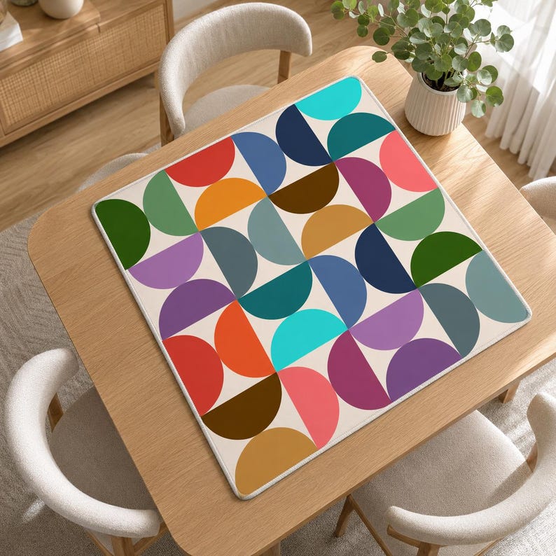 Pu&ograve; includere: Un tappetino quadrato con un motivo geometrico ripetitivo di semicerchi multicolori disposti a griglia. Il design include sfumature di rosso, arancione, giallo, verde, blu, viola e marrone su uno sfondo color crema chiaro.
