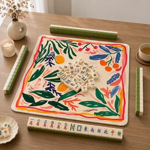 May include: A mahjong set featuring a square play mat with a cream background and a colorful botanical print of green leaves, orange fruits, and red berries. The set includes white tiles with various symbols and green backs, arranged on a wooden surface.
