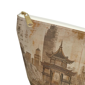 May include: A beige zippered pouch featuring a sepia-toned illustration of a cityscape with a pagoda and skyscrapers. The pouch has a gold-colored zipper and a cream-colored interior lining. The design gives the pouch a vintage aesthetic.