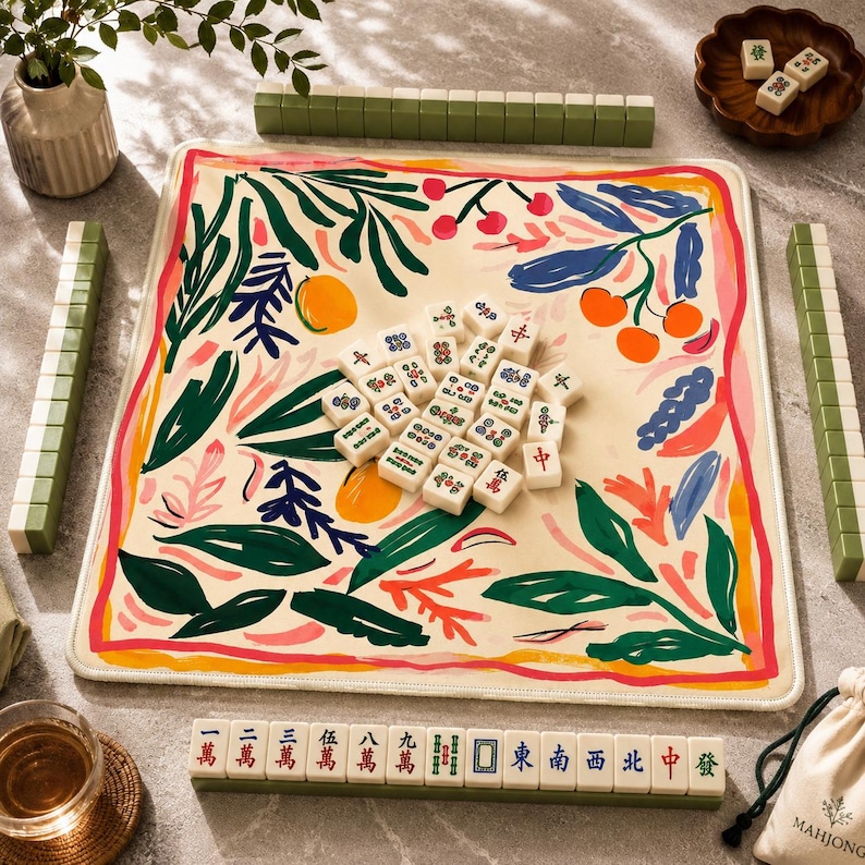 May include: A square mahjong mat with a cream background featuring a colorful botanical print of green leaves, orange fruits, and pink accents. A cluster of white mahjong tiles sits in the center, with additional tiles arranged in racks and a small bowl.