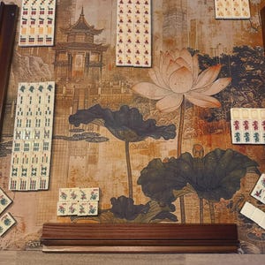 May include: A Mahjong set with white tiles featuring various red, green, and black symbols, characters, and floral designs arranged on a game mat. The mat displays a printed illustration of a traditional pagoda, a large lotus flower, and lily pads.