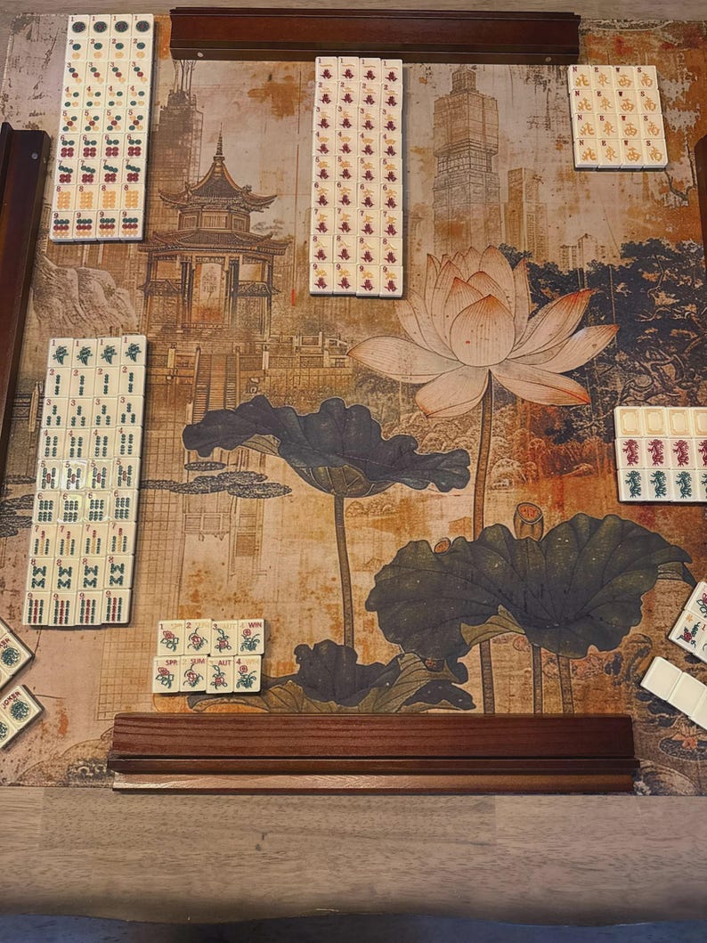 May include: A Mahjong set with white tiles featuring various red, green, and black symbols, characters, and floral designs arranged on a game mat. The mat displays a printed illustration of a traditional pagoda, a large lotus flower, and lily pads.