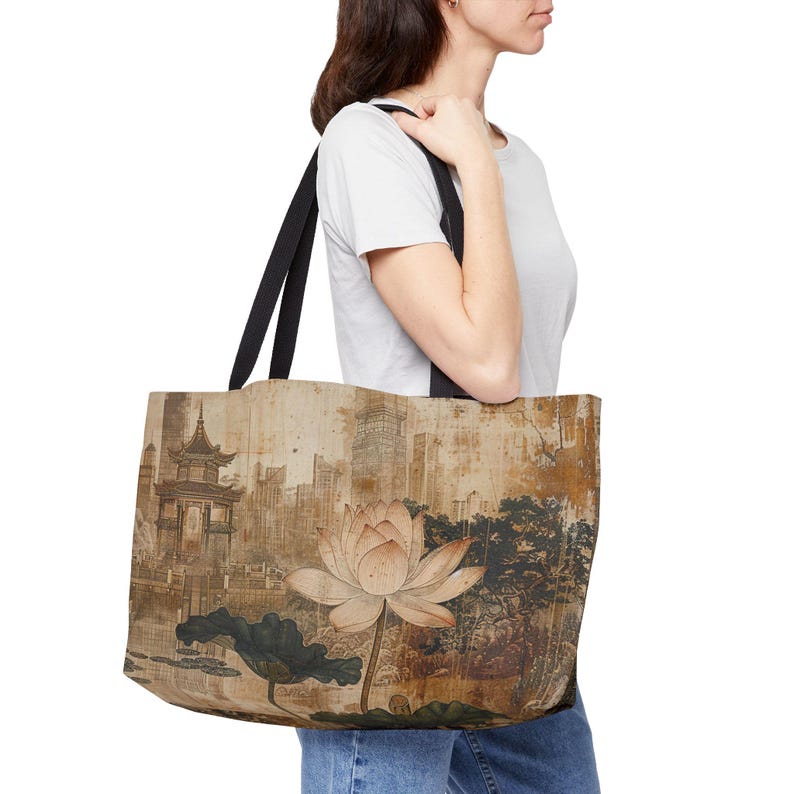 May include: A beige tote bag featuring a watercolor painting of a lotus flower, a traditional pagoda, and a cityscape. The bag has black straps and is carried by a person wearing a white t-shirt and blue jeans.