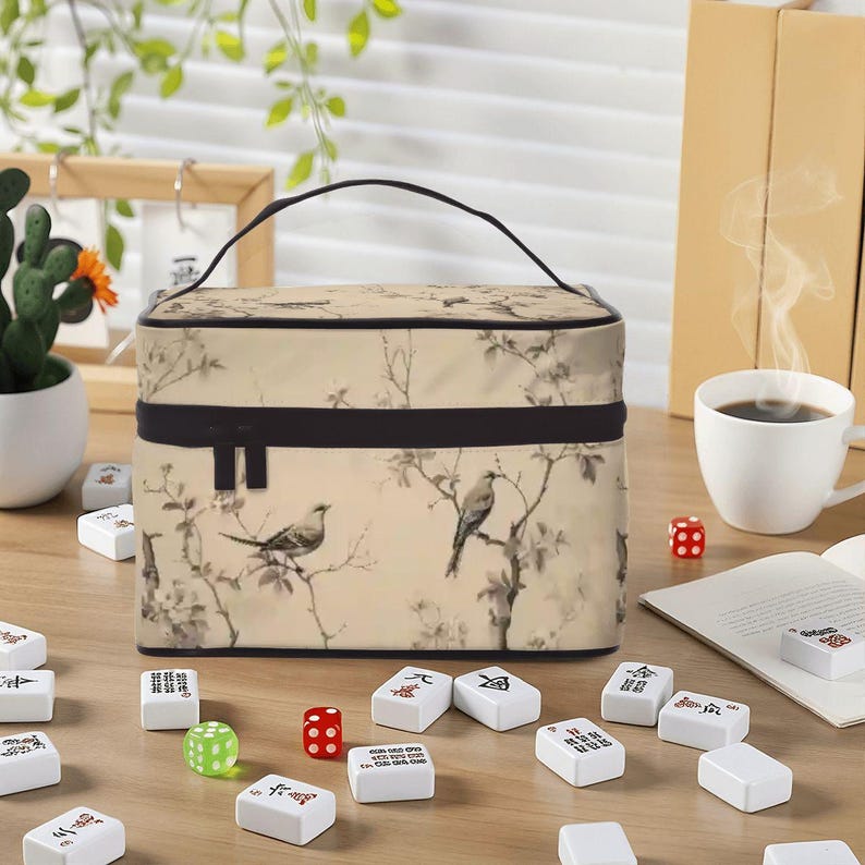 Op de afbeelding: Een beige make-uptas met een zwart handvat en rits, met een vogel- en bloemenprint. Mahjong-tegels, dobbelstenen en een kopje koffie omringen de tas, wat wijst op een spel- of reisthema.