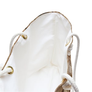 May include: A white tote bag with a light brown map-like pattern on the exterior. The bag has white rope handles and gold-colored grommets. The interior is white.