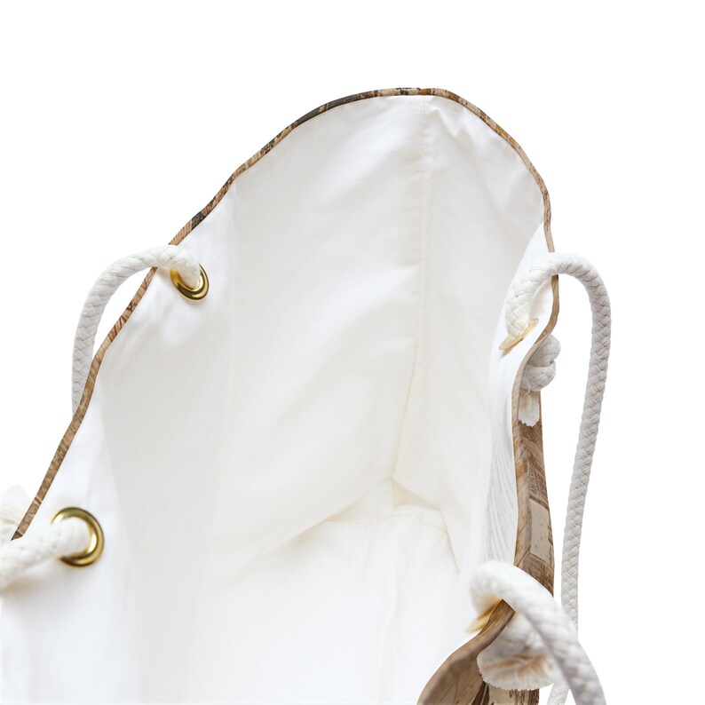 May include: A white tote bag with a light brown map-like pattern on the exterior. The bag has white rope handles and gold-colored grommets. The interior is white.