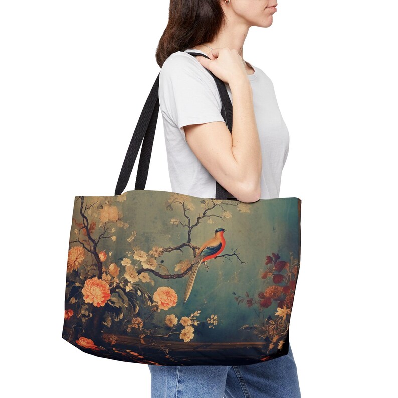 May include: A large tote bag with black straps featuring a vintage-style painting of a bird perched on a branch with flowers. The artwork has a teal background with orange, red, and yellow floral accents.
