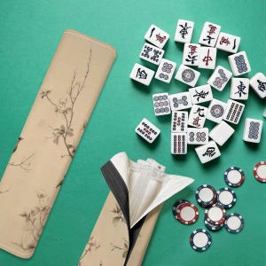 Op de afbeelding: Een beige Mahjong-tegelkoffer met een bloemenpatroon, open om de tegels te laten zien. De tegels zijn wit met zwarte en rode markeringen. Pokermunten met rode, witte en blauwe randen zijn ook zichtbaar.