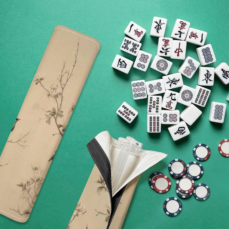 Op de afbeelding: Een beige Mahjong-tegelkoffer met een bloemenpatroon, open om de tegels te laten zien. De tegels zijn wit met zwarte en rode markeringen. Pokermunten met rode, witte en blauwe randen zijn ook zichtbaar.