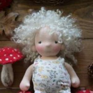 May include: A handmade cloth doll with a whimsical design. The doll has curly white hair, rosy cheeks, and is wearing a floral print romper with white lace trim. Two red and white mushroom decorations are on either side of the doll.