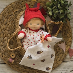 Puede incluir: Una muñeca hecha a mano con un gorro de punto rojo y un mono blanco y rojo con estampado de fresas está tumbada en una cesta tejida. La muñeca sostiene una muñeca pequeña en su mano. La muñeca está rodeada de una manta blanca con estampado de erizo.