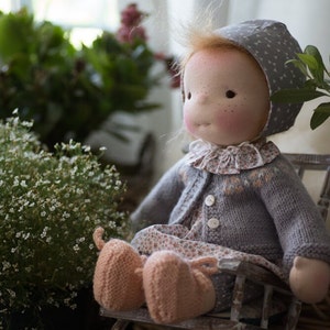 May include: A handmade doll with a soft fabric face, rosy cheeks, and freckles. The doll wears a gray knit sweater, a floral print dress, and a matching bonnet. It sits on a small wooden chair, with peach-colored knitted shoes.