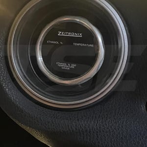 Puede incluir: Un indicador circular Zeitronix con un bisel plateado y una esfera negra. La pantalla presenta el texto Zeitronix, Ethanol %, Temperature y Ethanol % and Temperature Gauge impreso en letras blancas sobre el fondo oscuro.