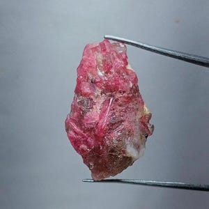 Może przedstawiać: Surowy, naturalny kamień szlachetny o żywym różowym odcieniu. Okaz minerału jest trzymany przez srebrną pęsetę na neutralnym szarym tle. Kamień ma krystaliczną strukturę i lekko przezroczysty wygląd.