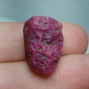 May include: A rough, pink ruby gemstone with a deep red hue. The gemstone has a natural, unpolished finish.