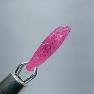 May include: A pink ruby crystal, with a pointed tip, is held in a metal clamp.