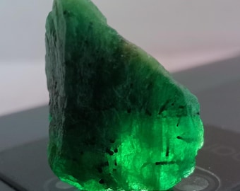 May include: A raw, uncut emerald gemstone with a vibrant green hue and translucent qualities. The stone features a jagged, irregular shape with visible dark mineral inclusions throughout its crystalline structure.
