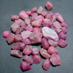 May include: A collection of pink ruby rough stones of various sizes and shapes. The stones are arranged in a pile on a gray surface.