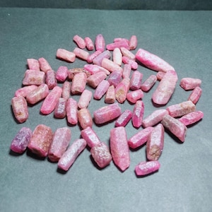 Puede incluir: Una colección de gemas de rubí en bruto, sin cortar, en varios tonos de rosa y rojo. Las gemas tienen forma irregular, con una textura rugosa y natural. Están dispersas sobre una superficie gris oscuro.