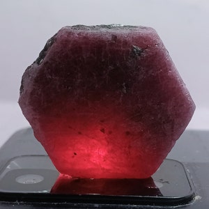 May include: A large, rough, dark red ruby gemstone with a hexagonal shape. The ruby is illuminated from below, highlighting its deep red colour.