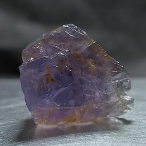 Puede incluir: Un espécimen de cristal crudo y translúcido de color púrpura con bordes irregulares y dentados y texturas superficiales naturales. La piedra muestra varios tonos de violeta claro y lavanda, con sutiles inclusiones marrones en su estructura.