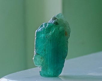 May include: A vibrant green emerald crystal with a rough, natural texture. The crystal is translucent and has a slightly pointed shape.