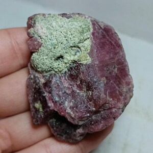 May include: A raw, uncut gemstone with a deep magenta hue, partially covered by a patch of light green mineral deposits. The rough texture and natural form of the stone are clearly visible, showcasing its organic beauty.