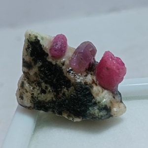 May include: A natural gemstone specimen featuring a cluster of vibrant, deep pink ruby crystals embedded in a matrix of speckled, light-colored rock with dark green and black inclusions. The rough ruby crystals have a raw, unpolished appearance.